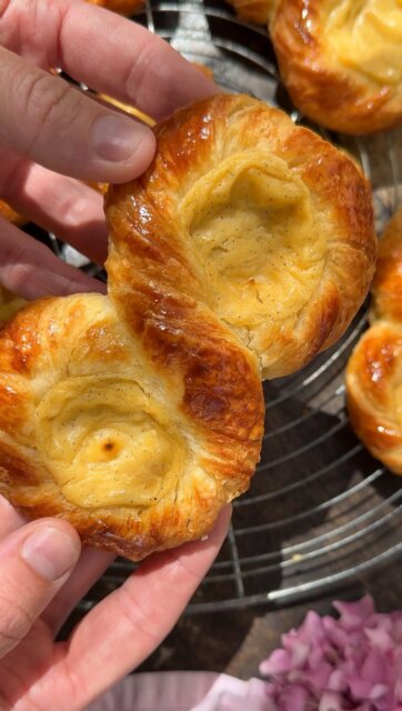 REZEPT: Unsere Vanilleknoten zum Nachbacken 🥨

Knuspriger Croissantteig trifft auf cremigen Vanillepudding – buttrig, luftig und einfach richtig lecker.

Croissantteig
20 g frische Hefe
100 g lauwarmes Wasser
420 g Mehl T45
52 g Zucker
10 g Salz
80 g Vollmilch
30 g kalte Butter
250 g kalte Butter zum Tourieren

Puddingmasse
400 g Vollmilch
100 g Sahne
1 Vanilleschote
70 g Zucker
40 g Maisstärke
50 g Vollei
80 g Eigelb

Außerdem
1 Ei zum Bestreichen

Läuterzucker
150 g Zucker
150 g Wasser

Die Hefe im Wasser auflösen und mit 100 g Mehl verrühren. Die Poolish 30 Minuten bei Raumtemperatur stehen lassen.

Mit den restlichen Zutaten, außer der Tourierbutter, etwa 7 Minuten langsam kneten. Den glatten Teig anschließend kalt stellen.

Die kalte Tourierbutter zwischen Backpapier mit dem Rollholz weich klopfen und zu einer flachen Platte ausrollen.

Tipp zum Tourieren:
Teig und Butter sollten kühl sein und vor allem eine ähnlich geschmeidige Konsistenz haben. Zu harte Butter bricht, zu weiche Butter tritt aus.

Den Teig etwa 8 mm dick ausrollen, die Butter einschlagen und drei einfache Touren geben. Wird der Teig zu weich, zwischendurch nochmals kühlen. Danach 1 Stunde kalt stellen.

Für den Pudding Milch, Sahne und Vanille aufkochen. Zucker und Stärke mischen und mit Vollei und Eigelb verrühren. Mit etwas heißer Milch angleichen, alles zurück in den Topf geben und unter Rühren eindicken lassen. Abgedeckt auskühlen, glatt rühren und in einen Spritzbeutel füllen.

Zucker und Wasser für den Läuterzucker einmal aufkochen.

Den Teig etwa 2,5 mm dick und ca. 55 × 15 cm groß ausrollen. In 1,5 cm breite Streifen schneiden, eindrehen und wie im Reel formen.

Bei ca. 25 °C etwa 2 Stunden gehen lassen. Mit Ei bestreichen und den Pudding in die Öffnungen spritzen.

Bei 195 °C Ober-/Unterhitze 15–20 Minuten goldbraun backen. Noch heiß mit Läuterzucker abglänzen.

Viel Freude beim Nachbacken! 💜✨